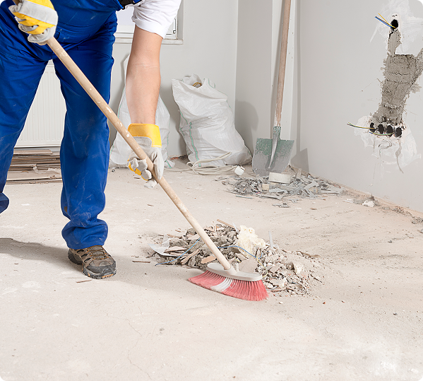 Person in blue overalls sweeping debris with a broom on a construction site floor. Tools and a white bag are visible in the background.